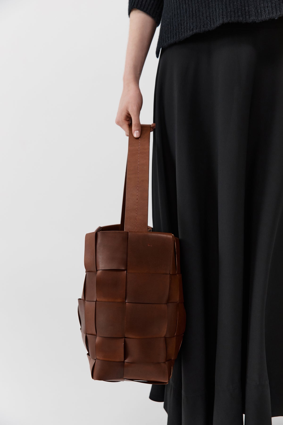 Brown woven handbag held by a person wearing a dark skirt against a white background